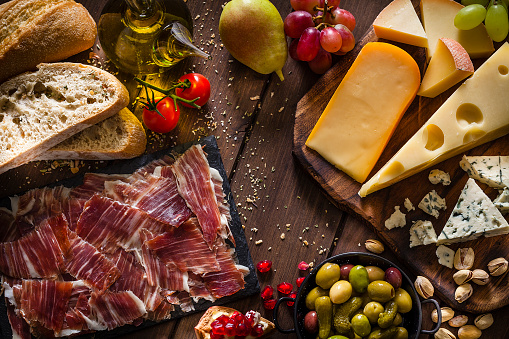 Typical Mediterranean food concepts: Top view of a rustic wooden table filled with delicious Mediterranean appetizer. The composition includes a cheese selection, Iberico ham slices, pears, cherry tomatoes, pomegranate, pickles, olive oil, grapes, olives, pistachio and bread. Predominant color is brown. Low key DSRL studio photo taken with Canon EOS 5D Mk II and Canon EF 100mm f/2.8L Macro IS USM.