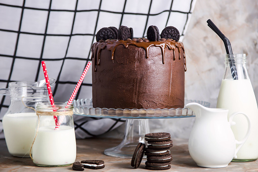 Chocolate cake with biscuits on a glass stand among the vessels with milk on a gray vintage background. Gourmet Pastry Concept