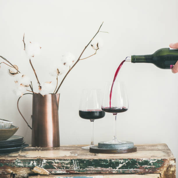 Red wine pouring from bottle in man's hand, square crop Red wine pouring from bottle in man's hand into wineglass over rustic kitchen countertop, white background behind, copy space, square crop. Wine shop, winery, bojole nouveau holiday concept liquor store counter photos stock pictures, royalty-free photos & images