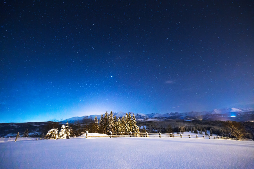 Starry winter night in the mountains. The sky is full of bright stars above the snow-covered valley and mountain peaks. Winter night landscape in blue tones.