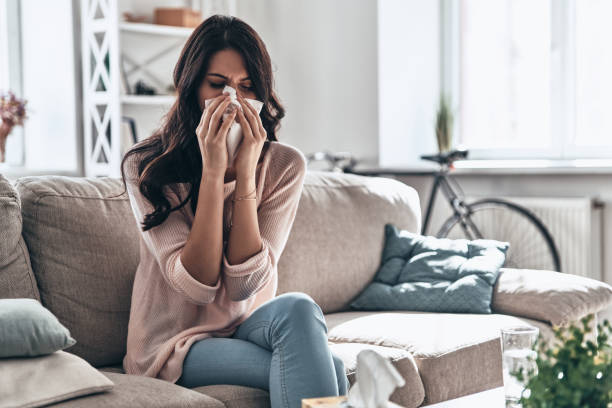 Cold and flu. Sick young woman blowing the nose using tissue paper while sitting on the sofa at home sneezing photos stock pictures, royalty-free photos & images