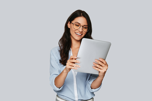 Attractive young woman working using digital tablet and smiling while standing against grey background