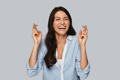 Attractive young woman crossing fingers and smiling while standing against grey background