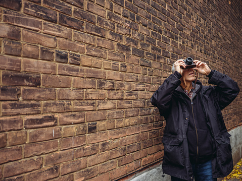 Senior man in fall outfit with retro digital camera standing in front of old brick wall.