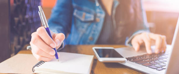 istock Woman hand is writing on a notepad with a pen and a phone with a laptop.Web banner. 1066999730