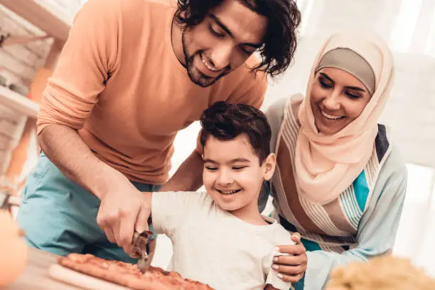 Happy Arabian Family Eating Pizza in Kitchen. Muslim Family. Smiling Boy. Young Arabian Woman. Modern Kitchen at Home. Man Using Kitchenware. Young Family. Wooden Table in Kitchen. Food on Table.