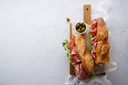 Top view of two fresh baguette sandwiches bahn-mi styled. Ham, sliced cheese, tomatoes and fresh lettuce on dark wooden cutting board on concrete background.
