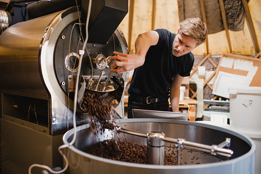 Male coffee shop employee using a coffee bean roasting machine and turning a dial to release the coffee beans.