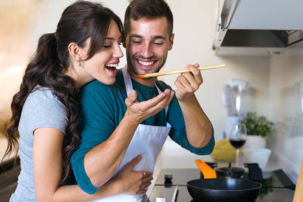 przystojny młody mężczyzna dając żonie, aby spróbować żywności przygotowuje w kuchni w domu. - cooking men caucasian togetherness zdjęcia i obrazy z banku zdjęć