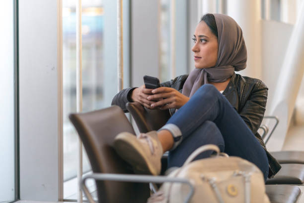 Waiting for a flight A muslim woman sits relaxed and comfortably in  set of chairs while she waits for her flight to arrive. She is looking out the window with her phone in her hand. airports canada stock pictures, royalty-free photos & images