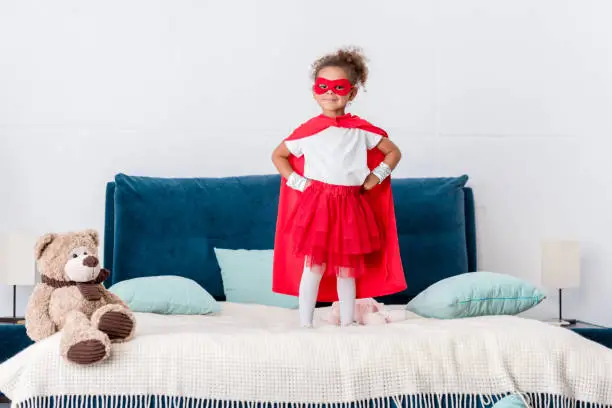 little african american kid in red superhero costume and mask with hands on hips standing on bed