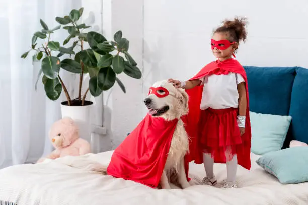 Cute little african american kid with dog in superhero costumes looking away while standing on the bed