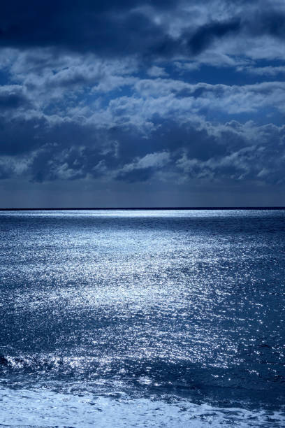 Sea and sky in the bottom half is a calm deep blue sea, on the horizon is a line of white shimmering glowing light from the moon in the upper half is the sky covered in fluffy clouds the image has a blue glow from the moonlight, vertical