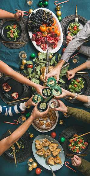 Company of friends gathering for Christmas or New Year party dinner at festive table. Flat-lay of hands holding glasses with drinks, feasting and celebrating holiday, top view, vertical composition