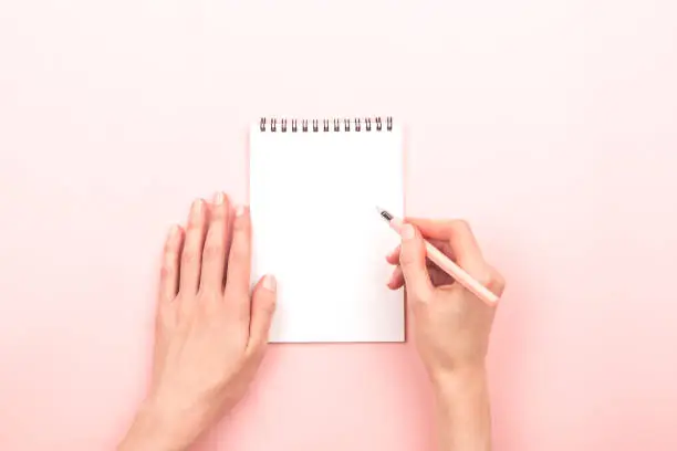 Woman hands are writing on notepad. Beautiful pastel pink background. Copy space for your text.
