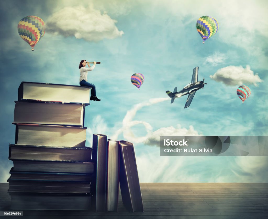 Adventure concept Surreal view as a young woman student stand on the top of a huge pile of books holding a spyglass looking far at horizon. Adventure concept, as airplane and hot air balloon fly out of the magic book. Book Stock Photo