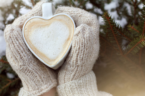hot mug of frothy cappuccino in hands dressed in mittens against the background of an evergreen tree in winter