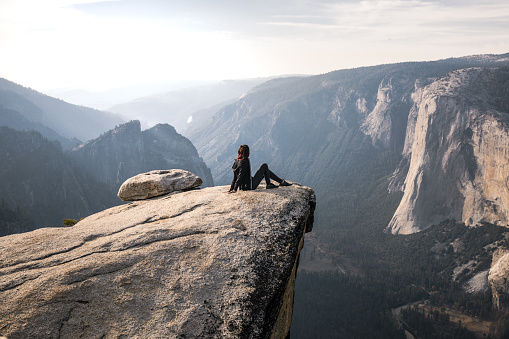 Beautiful mixed race brunette young woman seated at the edge of  a cliff looking out over Yosemite Valley. Seated leaning back on her hands. Excitement, inspiring.