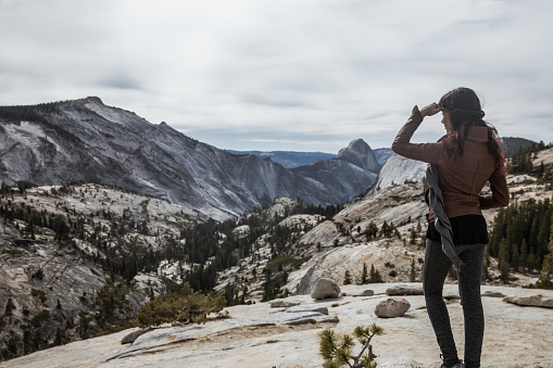 Fit young mixed race woman in Yosemite National Park gazing out at the valley. Back turned to camera with face partially showing and hand covering face from sun. Wearing brown leather jacket and hiking clothes.