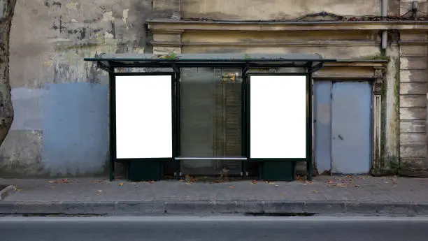 Photo of Two vertical blank white billboards at bus stop on old city street. In the background buildings and trees. Mock up. Poster on street next to roadway. Sunny summer day.