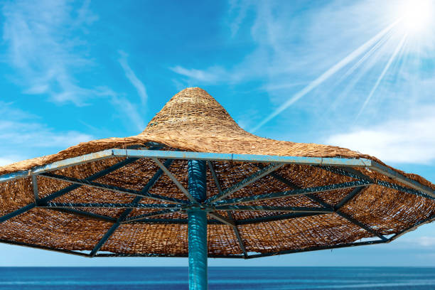 Close-up of a straw beach umbrella on a blue sky with sea and sun rays