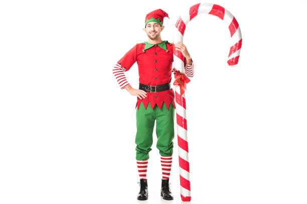 sonriente hombre de navidad traje de elf con la mano en las caderas junto a big candy cane aislado en blanco - elfo fotografías e imágenes de stock