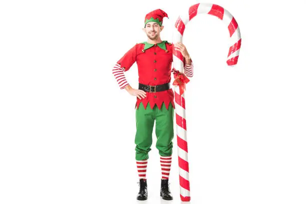 smiling man in christmas elf costume with hand on hips standing near big candy cane isolated on white
