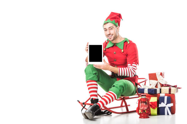 man in christmas elf costume sitting on sleigh and holding tablet with blank screen isolated on white man in christmas elf costume sitting on sleigh and holding tablet with blank screen isolated on white elf sitting stock pictures, royalty-free photos & images