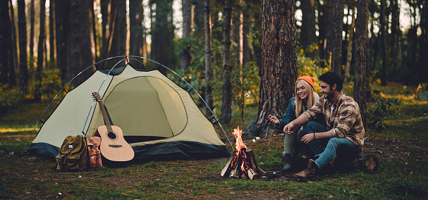 Young couple of tourists are exploring new places. Attractive woman and handsome man are spending time together on nature. Sitting near bonfire and touristic tent in forest and making marshmallow.