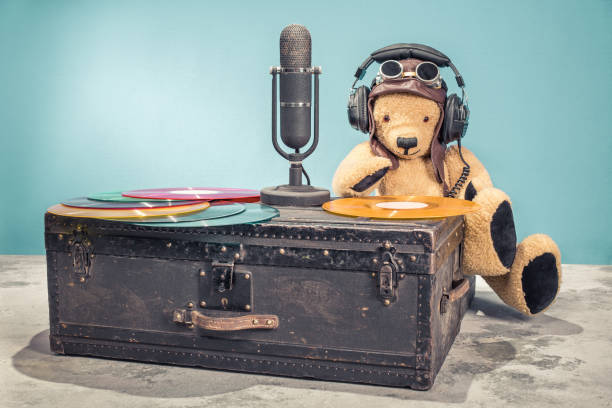istock Retro DJ Teddy Bear toy with leather aviator's hat, headphones and goggles and studio microphone, vinyl disk records on old aged classic travel trunk. Vintage style filtered photo 1067401846