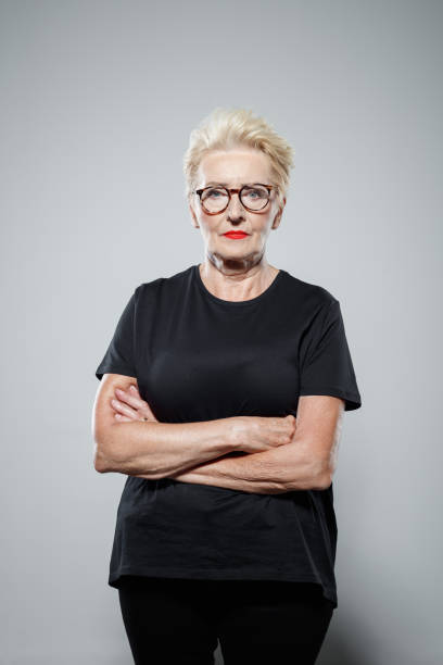 Portrait of confident senior woman Powerful senior woman wearing black clothes, standing with arms crossed against grey background, staring at camera. Studio shot. unconventional wisdom stock pictures, royalty-free photos & images