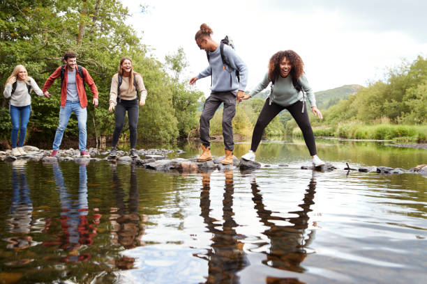 multi ethnischen gruppe von fünf junge erwachsene freunde lachen, als sie auf felsen, um während einer wanderung einen bach überqueren balancieren - couple young adult african descent multi ethnic group stock-fotos und bilder