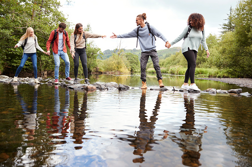 Young adult friends reaching to help each other cross a stream balancing on stones during a hike