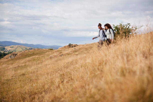 um casal adulto jovem raça mista sorrir enquanto atravessando uma descida de campo durante uma caminhada na montanha - adventure african ethnicity rural scene day - fotografias e filmes do acervo