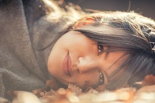 Beautiful mid adult woman enjoying wonderful day in nature. She lying down on ground covered with autumn leaves, wears gray sweater and she is absolutely gorgeous