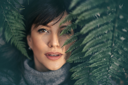 Beautiful mid adult woman enjoying wonderful day in nature. She is looking at camera from the green leaves of fern, while wears a gray sweater and looks amazing.