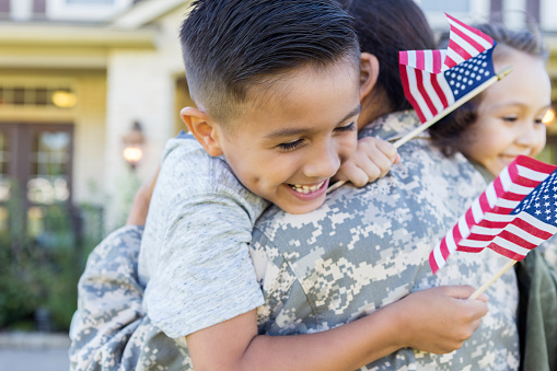 Smiling little boy and girl give their soldier mom a big hug on her return home from service. The children are holding small US flags.