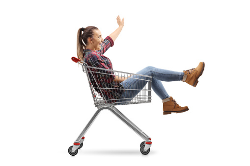 Young female sitting inside a shopping cart and waving isolated on white background
