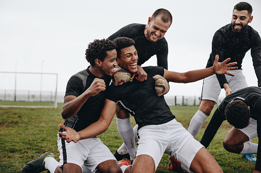 Football players celebrating success on the field. Happy footballer sitting on his knees with open arms after scoring a goal being cheered by his teammates.