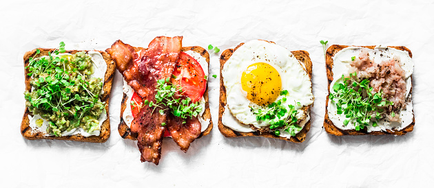 Variety of sandwiches for breakfast, snack, appetizers - avocado puree, fried egg, tomatoes, bacon, cream cheese, smoked mackerel grilled whole grain bread sandwiches. On a light background, banner view