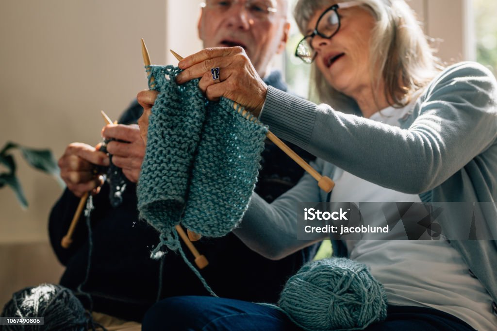 Senior couple knitting wool at home Senior woman teaching her husband the art of knitting woollen clothes. Senior man learning to knit woollen clothes from his wife sitting at home. Knitting Stock Photo