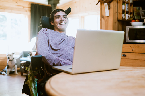 A cheerful young adult man with cerebral palsy researches something on his laptop, using voice recognition to aid in his studies.