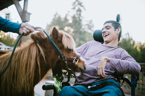 A cheerful young adult man with cerebral palsy spends time with a pony, used as a therapy animal.