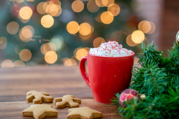 A red mug with whipped cream topped with red sprinkles on a wooden table. There are star-shaped gingerbread cookies around the mug and green fir branches, and a red bauble. A Christmas tree is in the background.