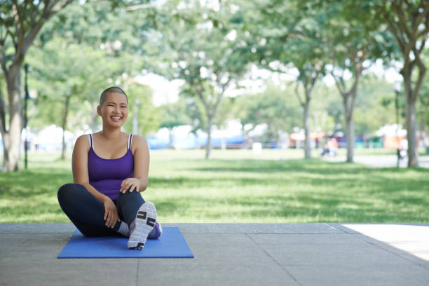 Active lifestyle Laughing bald young Asian woman exercising on yoga mat in city park survival stock pictures, royalty-free photos & images