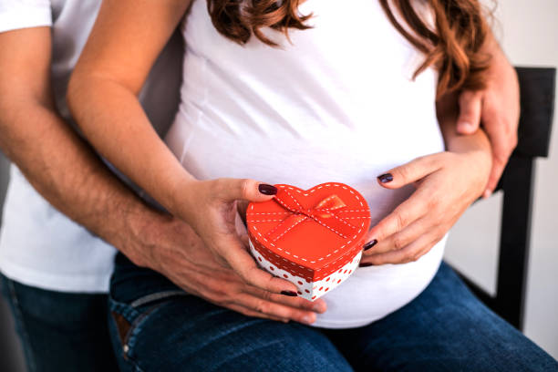 Pregnant couple hug and hold pregnant belly. Happy parents hug and hold pregnant belly. Woman hold a gift from her husband. They are happy to celebrate Valentine's Day together. cute pregnant woman with gift stock pictures, royalty-free photos & images