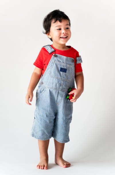 Young Caucasian girl toddler wearing denim overalls and black shirt raising arms happily. Child standing barefoot against curtain with warm lights creating cozy atmosphere.