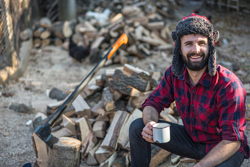 Young bearded lumberman sitting on the wood logs with a cup of coffee