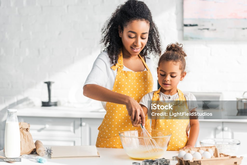 madre afroamericana e hija preparar la masa y batir huevos en cocina - Foto de stock de Cocinar libre de derechos