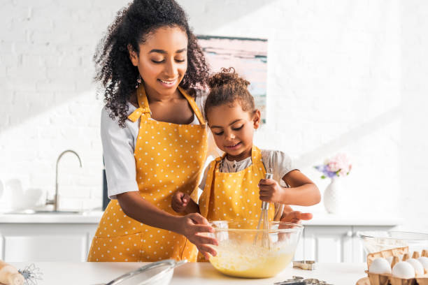 uśmiechnięta matka afroamerykanina pomaga córce przygotować i ubijać ciasto w kuchni - preschooler child offspring purity zdjęcia i obrazy z banku zdjęć